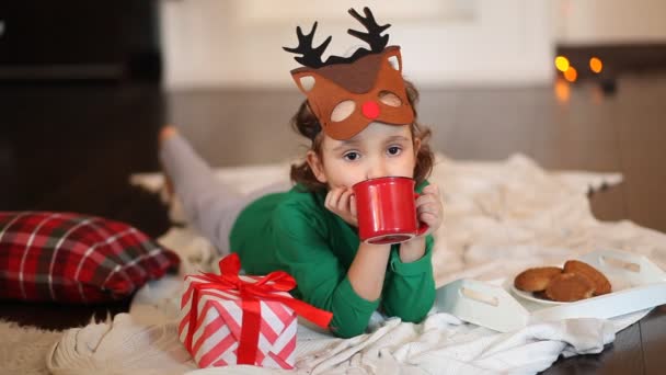 Dziewczynka w zabawnej masce jelenia i świątecznej piżamie leży na kratce jedząc ciasteczka i pijąc kakao — Wideo stockowe