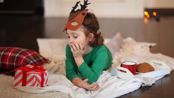 Niña linda en divertida máscara de ciervo y pijama de Navidad se encuentran en la manta con regalo, galletas y cacao. — Vídeos de Stock