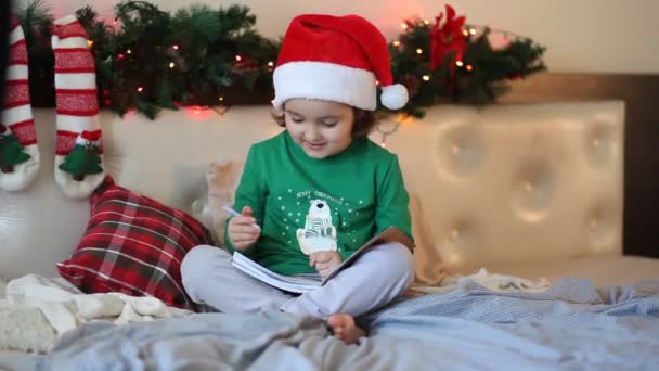 Zabawna dziewczyna w kapeluszu Mikołaja pisząc list do Świętego Mikołaja siedzącego na łóżku — Wideo stockowe