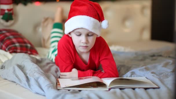 Niño con gorra de Santa Claus y pijama de Navidad leyendo un libro. — Vídeo de stock