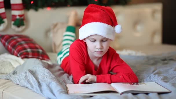 Παιδί με καπέλο και Χριστουγεννιάτικες πιτζάμες διαβάζει ένα βιβλίο.. — Αρχείο Βίντεο