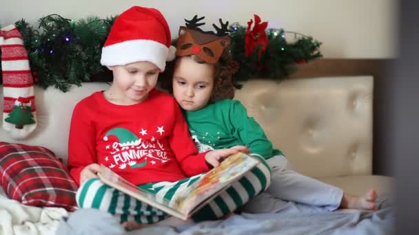 Little cute girl w zabawnej masce jelenia i chłopca w Santa cap i Boże Narodzenie piżamy czytając książkę. — Wideo stockowe