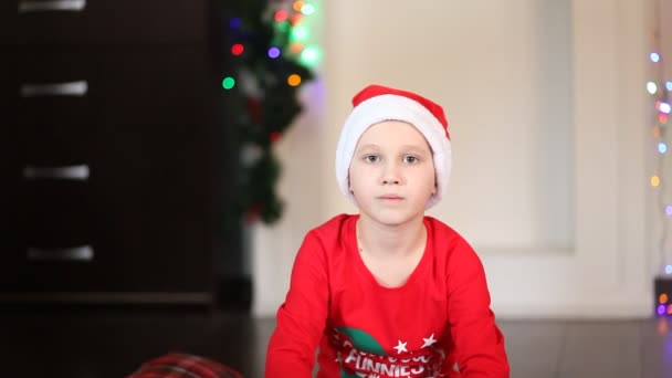 Little cute boy w zabawny Santa czapki i piżamy Boże Narodzenie siedzi na kocu i dostać pudełko prezentów — Wideo stockowe