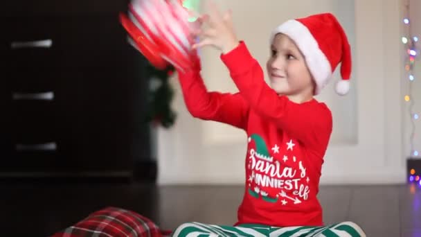 Little cute boy w zabawny Santa czapki i piżamy Boże Narodzenie siedzi na kocu i dostać pudełko prezentów — Wideo stockowe