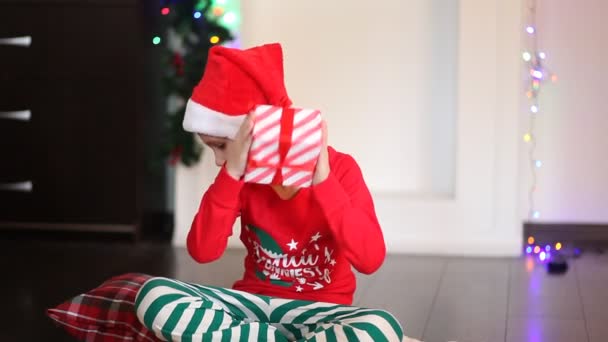 Kis aranyos fiú vicces Mikulás sapka és karácsonyi pizsama ül a takarón, és nézd meg az ajándék doboz — Stock videók