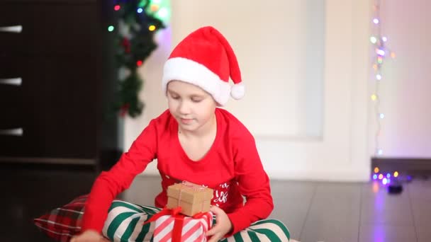 Little cute boy w zabawnej czapce Santa i piżamy Boże Narodzenie siedzi na kocu i przygotowuje prezenty dla bliskich — Wideo stockowe