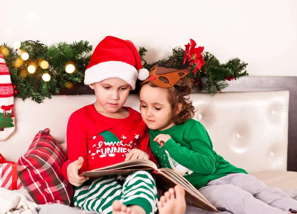 Kleines nettes Mädchen mit lustiger Hirschmaske und Junge mit Nikolausmütze und Weihnachtsschlafanzug lesen ein Buch. Stockbild