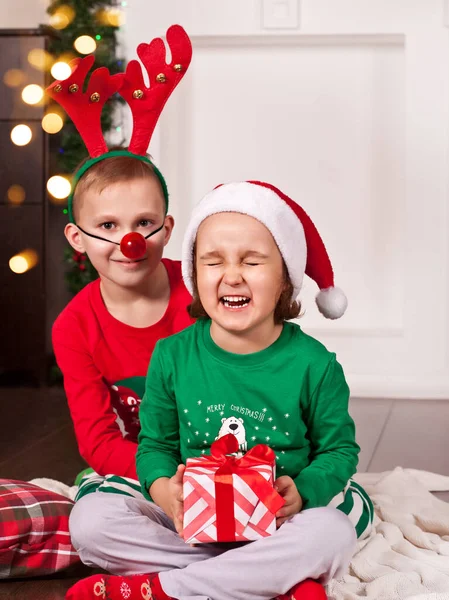 Feliz niña sonriente y niño en Santa Gorra y máscara de ciervo sentado en la manta en pijama de Navidad con caja de regalo. Imagen De Stock