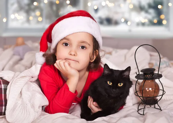 Kleines Mädchen in lustiger Weihnachtsmütze und Weihnachtsschlafanzug mit Lieblingstier schwarze Katze lizenzfreie Stockbilder