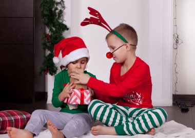 Noel Baba şapkalı ve geyik maskeli mutlu küçük kız ve oğlan hediye kutusuyla Noel pijamalarıyla battaniyenin üzerinde oturuyor..