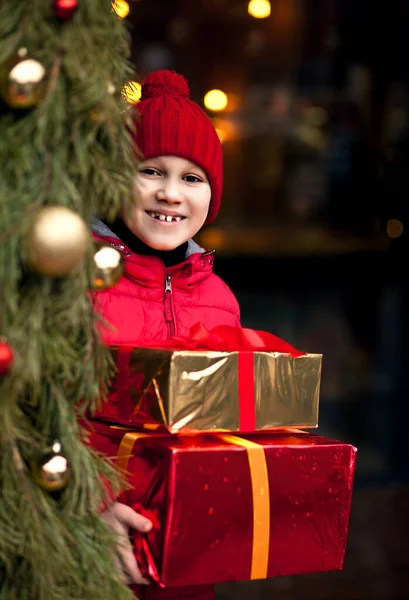 Chłopiec w zimie ubrania stojące na zewnątrz z ozdobioną choinką, trzymając pudełka prezentów — Zdjęcie stockowe