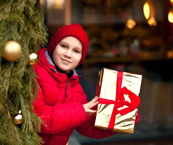 Хлопчик у зимовому одязі, що стоїть надворі з прикрашеною ялинкою, тримає подарункову коробку . — стокове фото