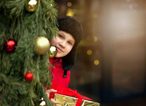 Chłopiec w zimie ubrania stojące na zewnątrz z ozdobioną choinką, trzymając pudełko prezentów. — Zdjęcie stockowe