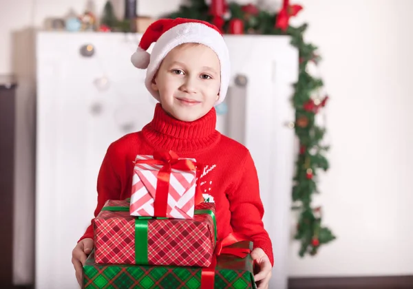 Хлопчик у в'язаному червоному светрі та шапочці Санти, що тримає різдвяні та новорічні подарункові коробки вдома . — стокове фото