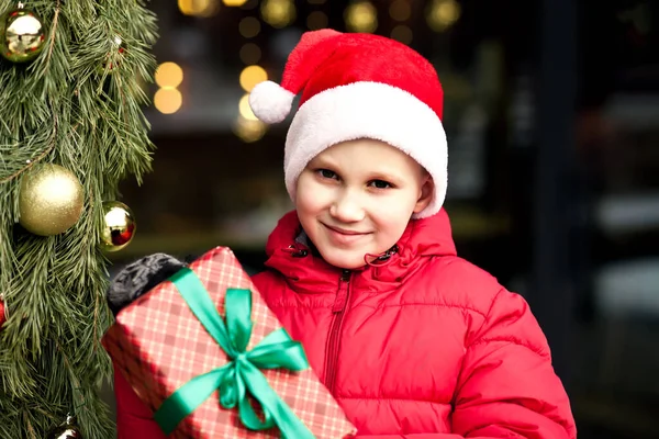 Chłopiec w zimowych ciuchach stojący na zewnątrz przy sklepie, trzymający pudełko z prezentami. Boże Narodzenie i Nowy Rok koncepcji zakupów. — Zdjęcie stockowe