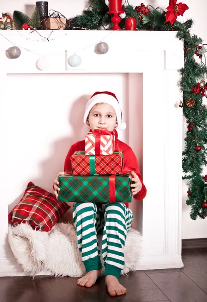 Boy kid in knitted red sweater and Santa cap holding Christmas and New Year gift boxes at home. Royalty Free Stock Images
