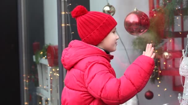 Αξιολάτρευτο παιδί κοιτάζει μέσα από το παράθυρο της βιτρίνας στη χριστουγεννιάτικη διακόσμηση στο κατάστημα — Αρχείο Βίντεο