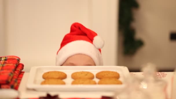 Kluk v Santa čepici se snaží ukrást sušenku ze stolu. — Stock video