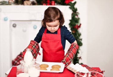 Little cute girl in red apron baking Christmas cookies at home.