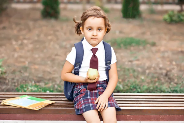 Üniformalı küçük tatlı kız bankta oturuyor ve elma yiyor. — Stok fotoğraf