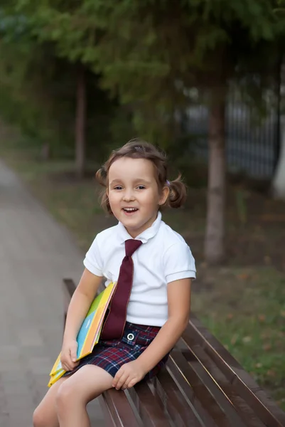 Маленька мила школярка в уніформі сидить на лавці і сміється — стокове фото