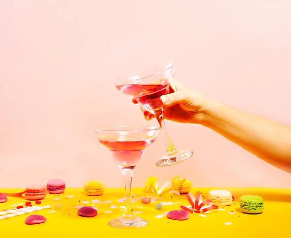 Ett glas rosa cocktail med hjärtgodis och makaroner. Födelsedagsfest eller Alla hjärtans dag koncept. Kvinnlig hand som håller ett glas. — Stockfoto