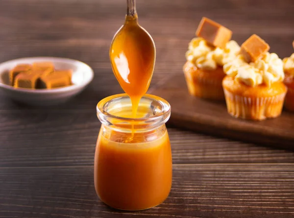 Lekker gezouten karamel met lepel uit glazen pot aan houten tafel en cupcakes op de achtergrond. — Stockfoto