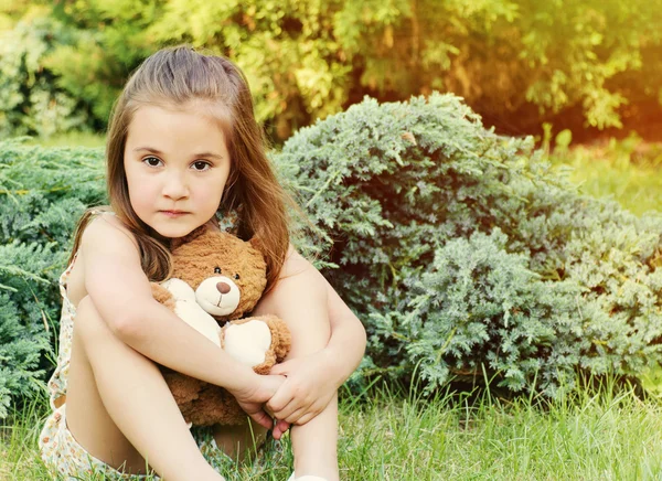 Красивая маленькая девочка, держащая игрушку и сидящая на улице на гр — стоковое фото