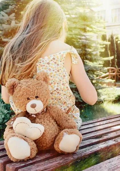 Gadis kecil duduk kembali dengan boneka beruang di bangku di bawah sinar matahari — Stok Foto