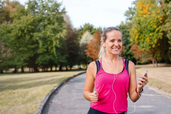 美丽而成熟的金发女子在阳光灿烂的一天在公园里奔跑 女跑步者慢跑时听音乐 健康生活方式概念 图库图片