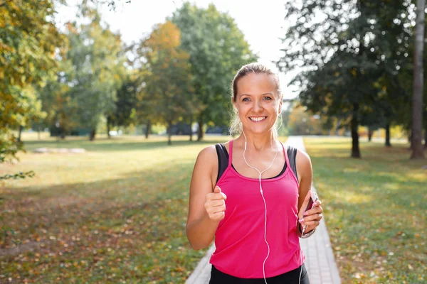 美丽而成熟的金发女子在阳光灿烂的一天在公园里奔跑 女跑步者慢跑时听音乐 健康生活方式概念 免版税图库照片