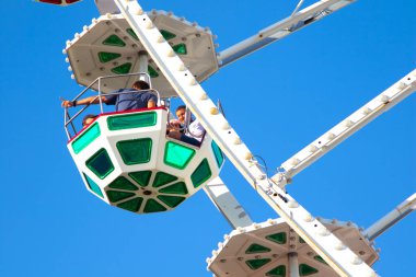 İnsanlar dönme dolaba biniyor. Mavi gökyüzüne karşı dönme dolap. Adam parmağını yana uzatıyor..