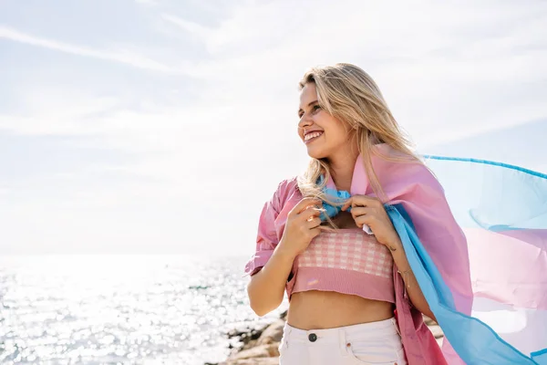 Woman standing on seashore on windy day holding transgender flag — Stock Photo, Image