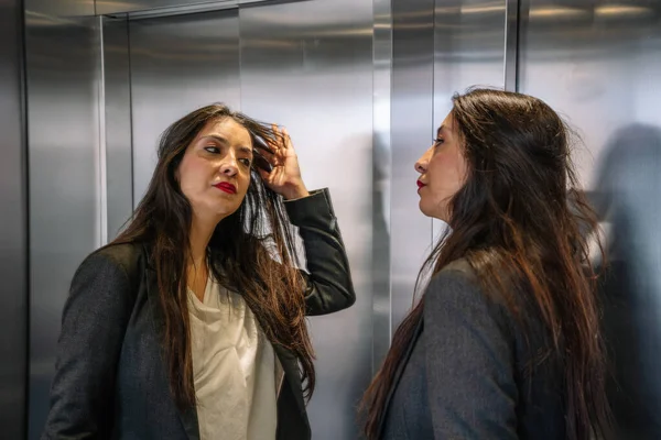 Bien habillé femme d'affaires hispanique toucher les cheveux dans l'ascenseur Image En Vente