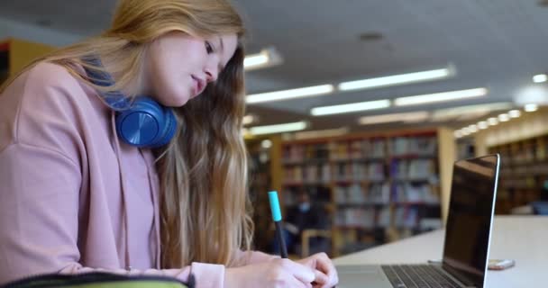 Kvinnlig student som sitter i biblioteket och gör läxor med hörlurar, bärbar dator framför sig — Stockvideo