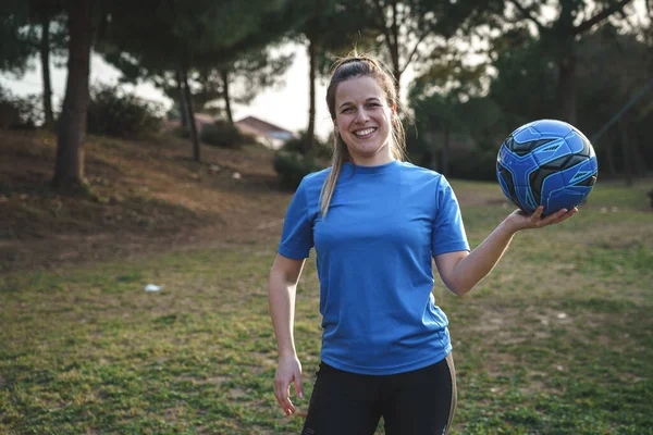 Üniformalı, at kuyruklu, top taşıyan ve parkta antrenman yaparken omzunun üzerinden kameraya bakan bayan futbolcu.