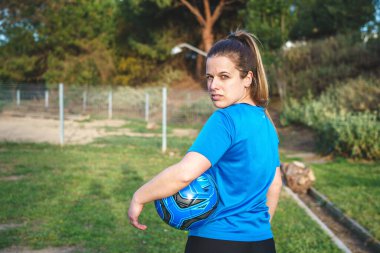 Üniformalı tam vücut özgüvenli sporcu el ele tutuşup, akşamları parkta futbol topuna basarken diğer tarafa bakıyor.