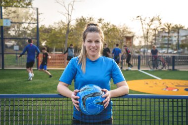 Üniformalı tam vücut özgüvenli sporcu el ele tutuşup, akşamları parkta futbol topuna basarken diğer tarafa bakıyor.