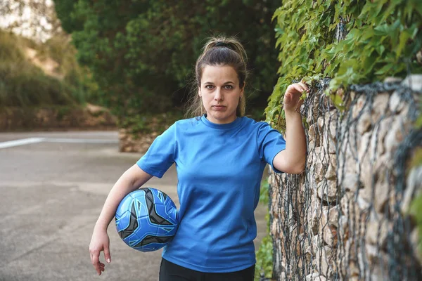 Determined young female soccer player with long ponytail in blue sportswear standing on path near lush green plants with ball in hand and looking at camera