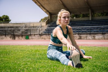 Full body female athlete touching foot and looking away while sitting on lawn during training on track and field stadium in summer