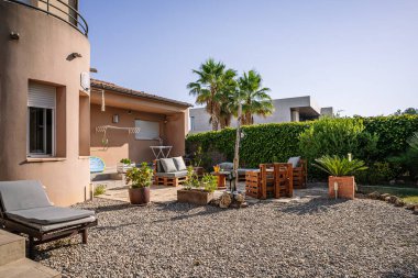 Comfortable benches and table placed in courtyard of cottage with green tropical trees under blue sky on sunny summer day