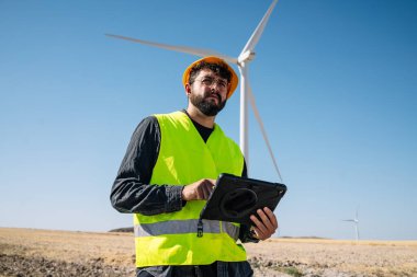 Low angle of serious bearded man in workwear and hardhat browsing data on tablet and looking away against windmill and cloudless blue sky during work on wind power station in daytime