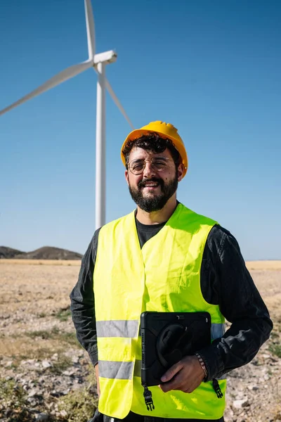 Positive bearded man in workwear and hardhat with tablet looking at camera with smile while standing near windmill against cloudless blue sky during work on wind power station