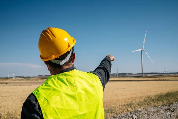 Anonymous man in vest and hardhat pointing at modern wind turbine against cloudless blue sky while standing in field on sunny summer day
