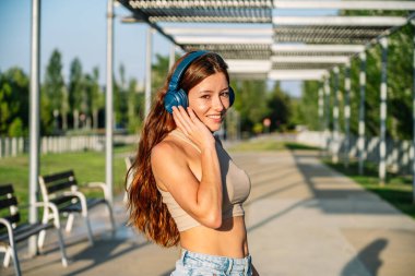 Side view of young content female meloman listening to music in wireless headphones while spending summer day in park and looking at camera
