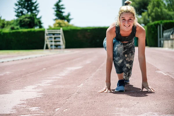 Full body happy female athlete in sportswear smiling and looking at camera while preparing to run from crouch start position during track and field training on sunny day