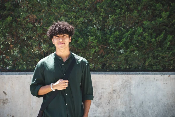 Beton sınırın ve caddede yeşil ağaçların yanında dikilirken kameraya bakan siyah saçlı neşeli genç bir erkek.