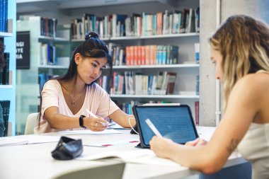 Üniversite kitaplarıyla kütüphanede ödev yaparken gazetelere yazan ve tablet kullanan odaklanmış çok ırklı kadın öğrenciler