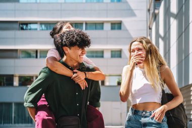 Neşeli, esmer, kıvırcık saçlı İspanyol adam, güneşli bir günde gülen kız arkadaşıyla sokakta yürürken kız arkadaşına sırtına biniyor.