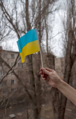 Bir adamın eli Ukrayna bayrağını gri bir gökyüzü, ağaç dalları ve sivil binanın arka planında tutuyor.